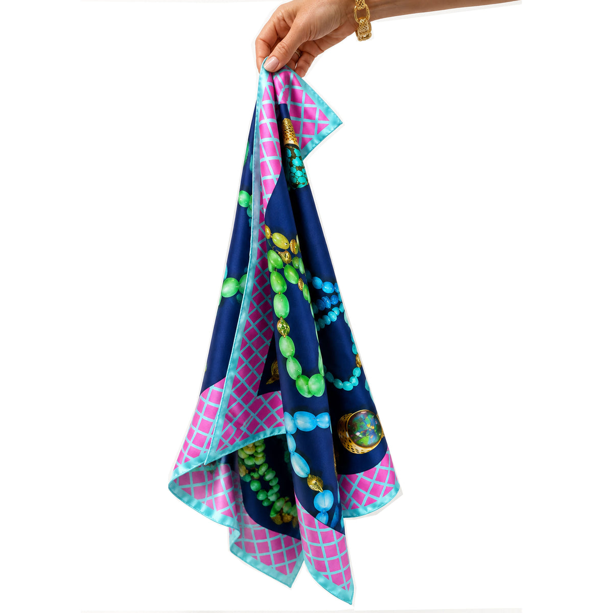 A hand holding a square silk scarf with a jewelry-themed print. The scarf features a navy blue background with green and turquoise beaded necklaces, pink lattice borders, and gold accents, set against a plain white background.