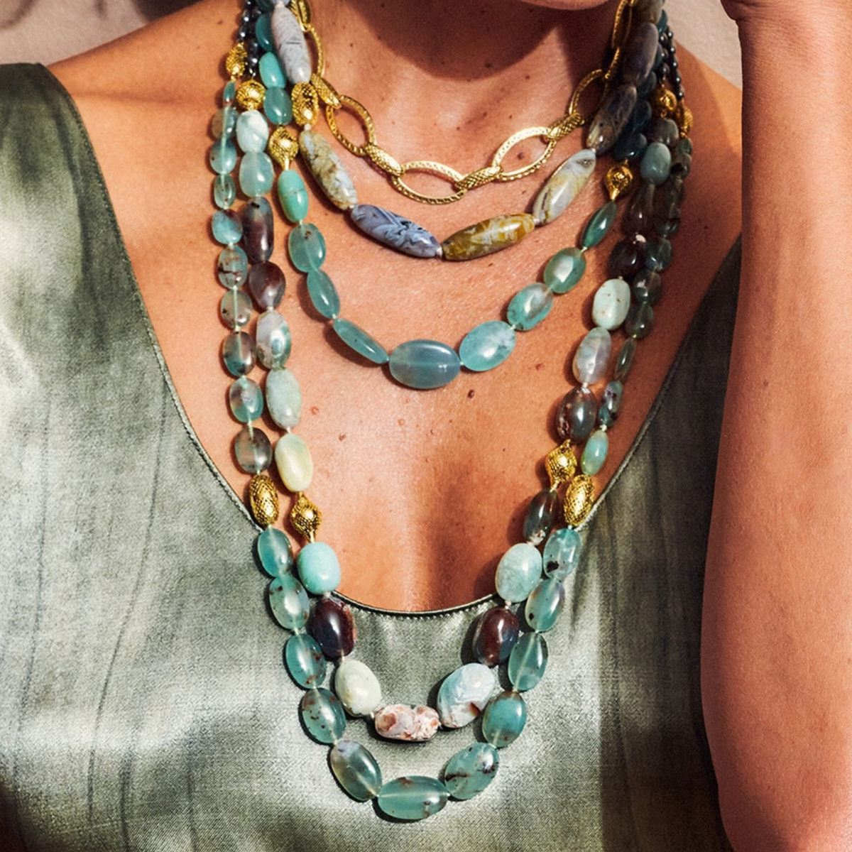 A close-up of a person wearing a layered necklace set. The necklace features multiple strands of polished turquoise and green stone beads, mixed with gold chain links and a few natural stone accents.