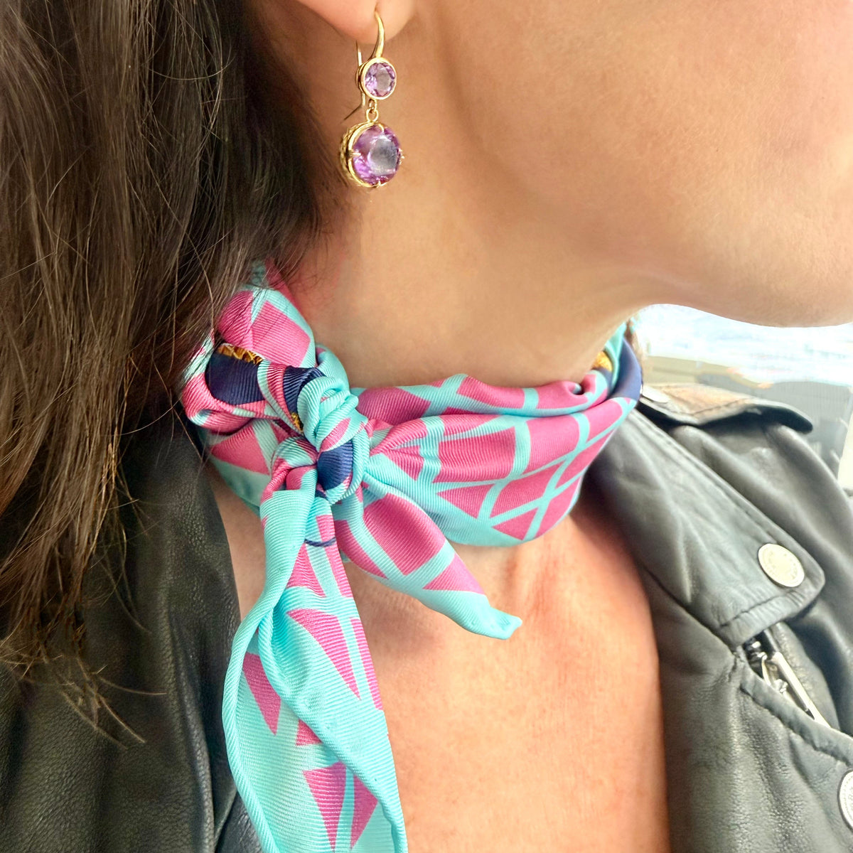 A close-up lifestyle shot of a woman wearing a vibrant pink and turquoise geometric pattern silk neck scarf tied in a knot at the base of her neck, paired with a black leather jacket.