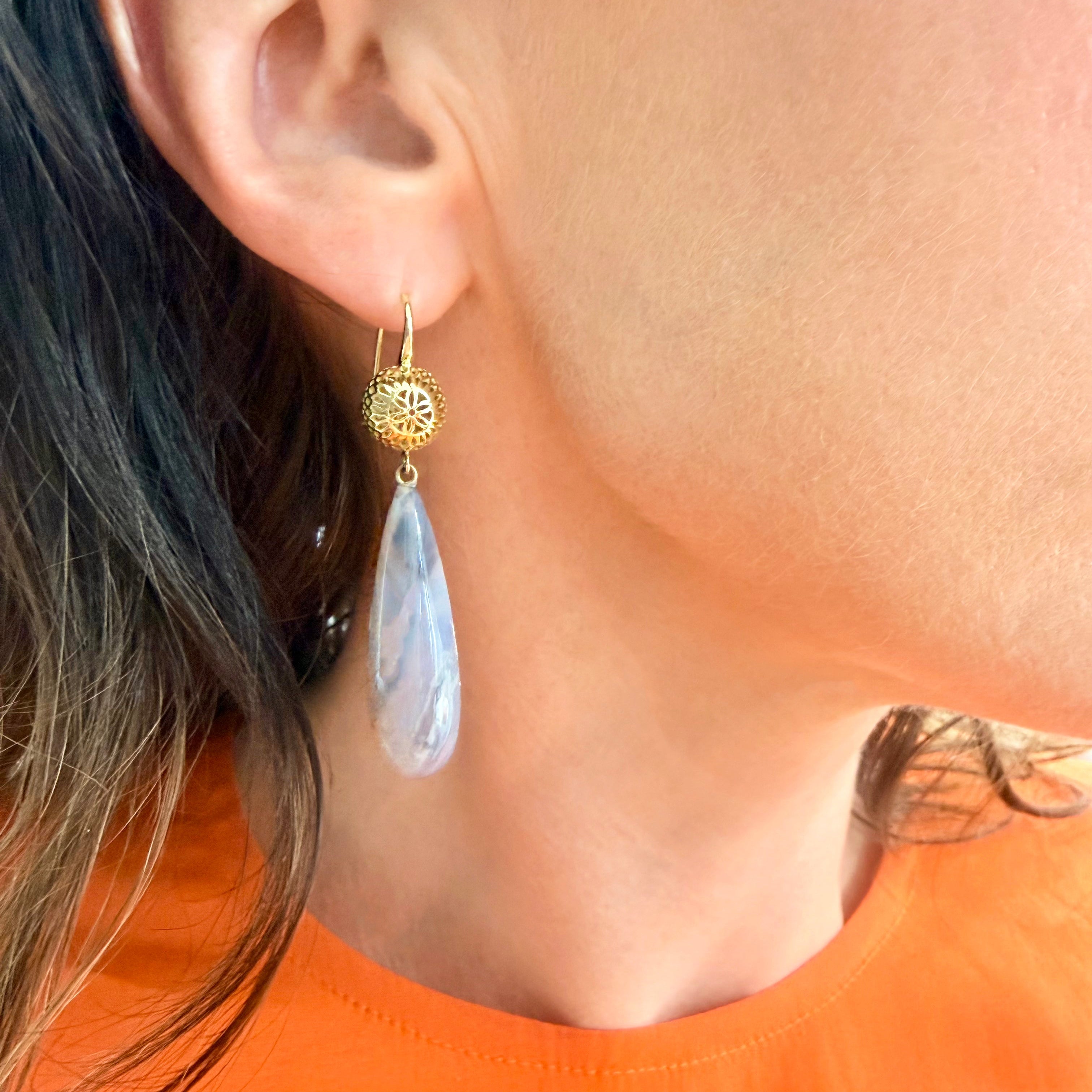 Close-up of an earring with a 18K YG Crownwork ball and boulder opal drop on a person wearing an orange top.