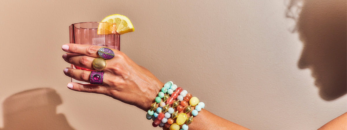A close-up of a hand holding a pink drink with a lemon slice, wearing colorful beaded bracelets and rings against a warm beige background with a soft shadow.