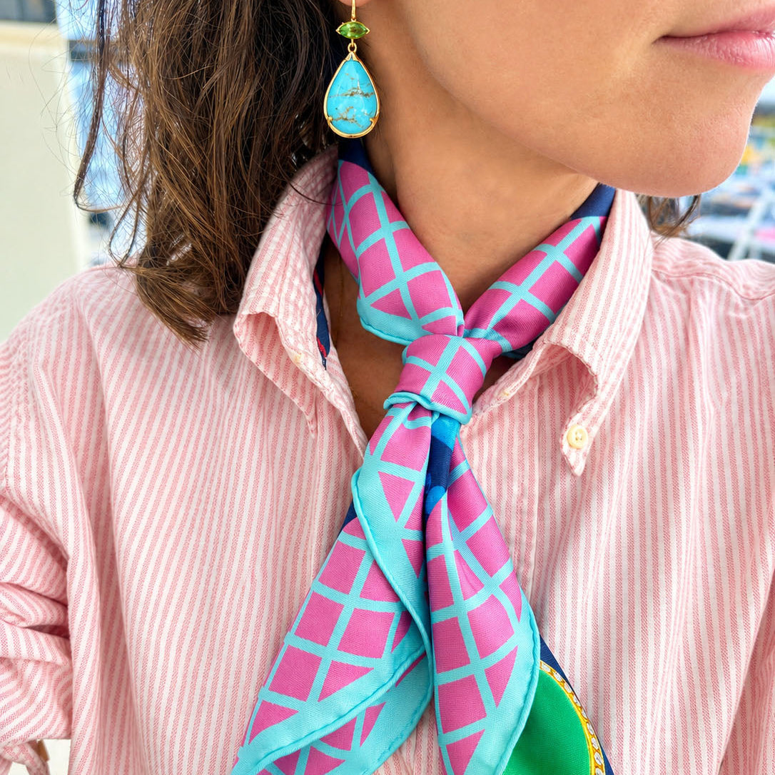 A close-up of a woman wearing a pink and turquoise patterned silk scarf tied around her neck, paired with a pink and white striped button-down shirt and a turquoise and tourmaline teardrop earring.