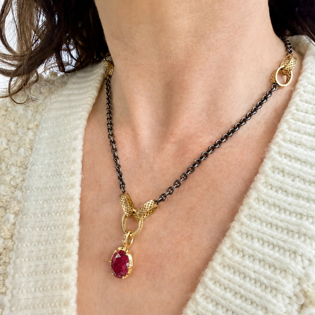 A close-up of a person wearing a gold & oxidized silver necklace with a crownwork  gold bale and a ruby oval pendant, paired with a cream-colored knit sweater.