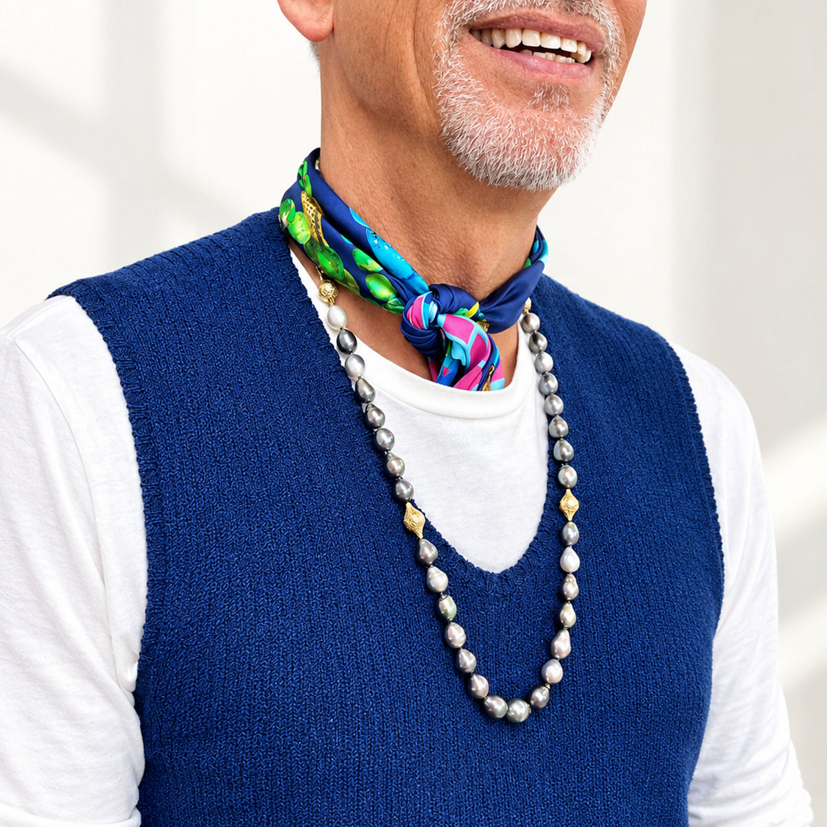 A close-up of a man wearing a blue knit sweater vest over a white long-sleeve shirt, accessorized with a colorful patterned silk neck scarf and a long silver pearl necklace.