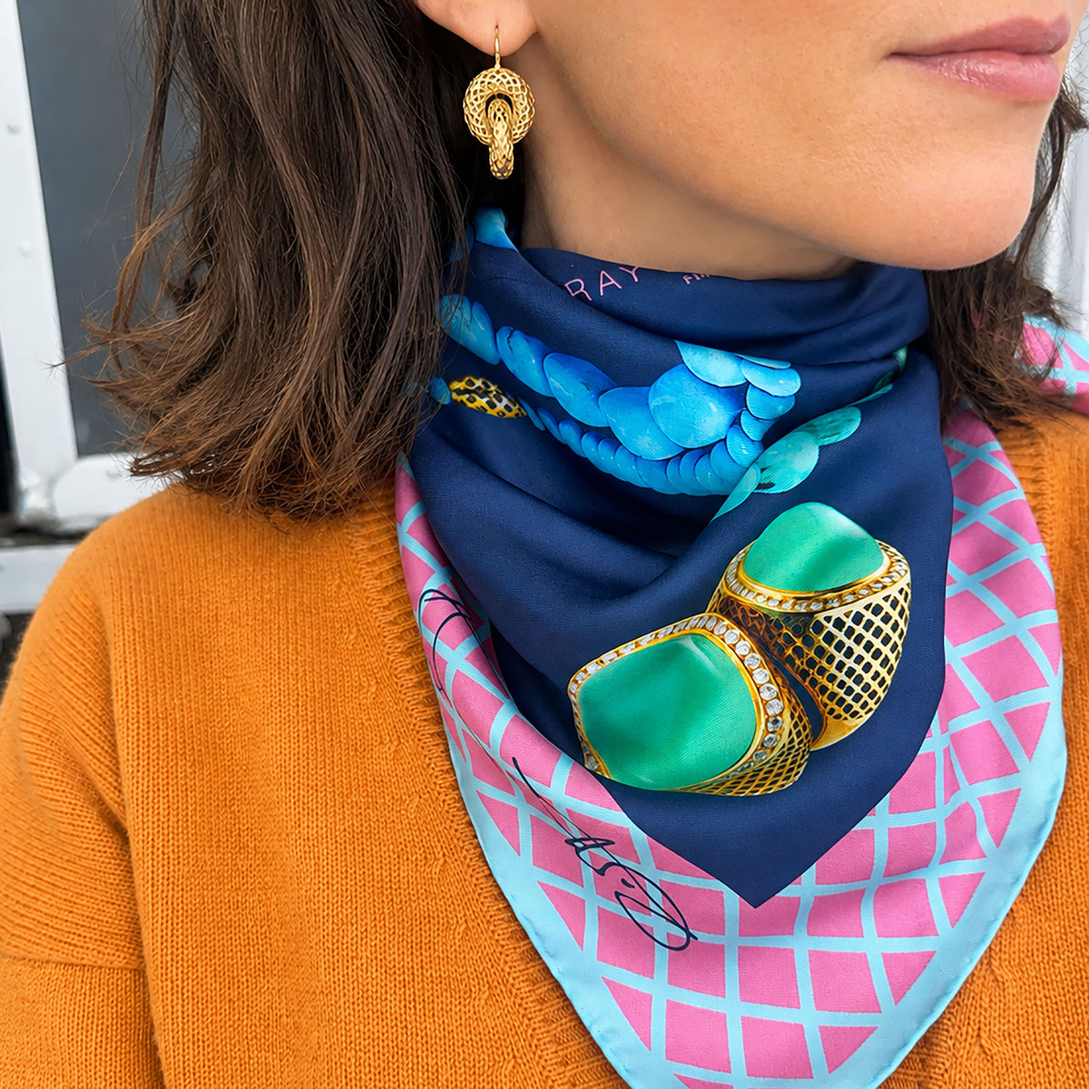 A close-up lifestyle shot of a woman wearing a navy blue silk scarf with a colorful print of turquoise necklaces, large green and gold rings, and a pink and light blue grid pattern on the reverse side. The model is also wearing an orange knit sweater and a gold crownwork earring.