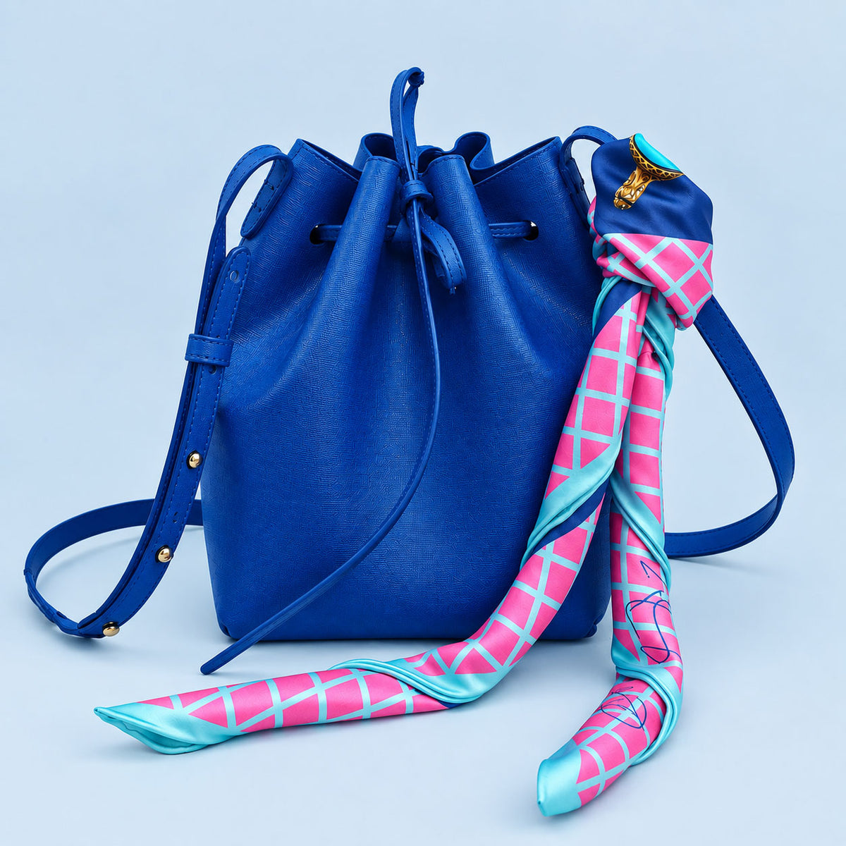 A royal blue leather bucket bag with a matching blue shoulder strap, styled with a pink and turquoise patterned silk scarf draped over the side against a light blue background.