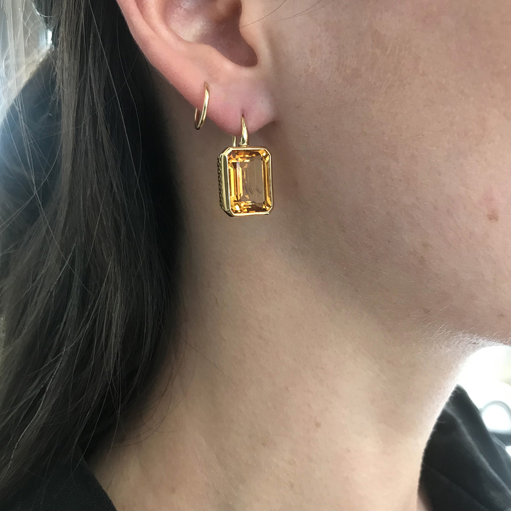 This is an image of a pari of emerald cut citrine bezel set earrings set in 18k Yellow Gold crownwork being worn to show how they hang